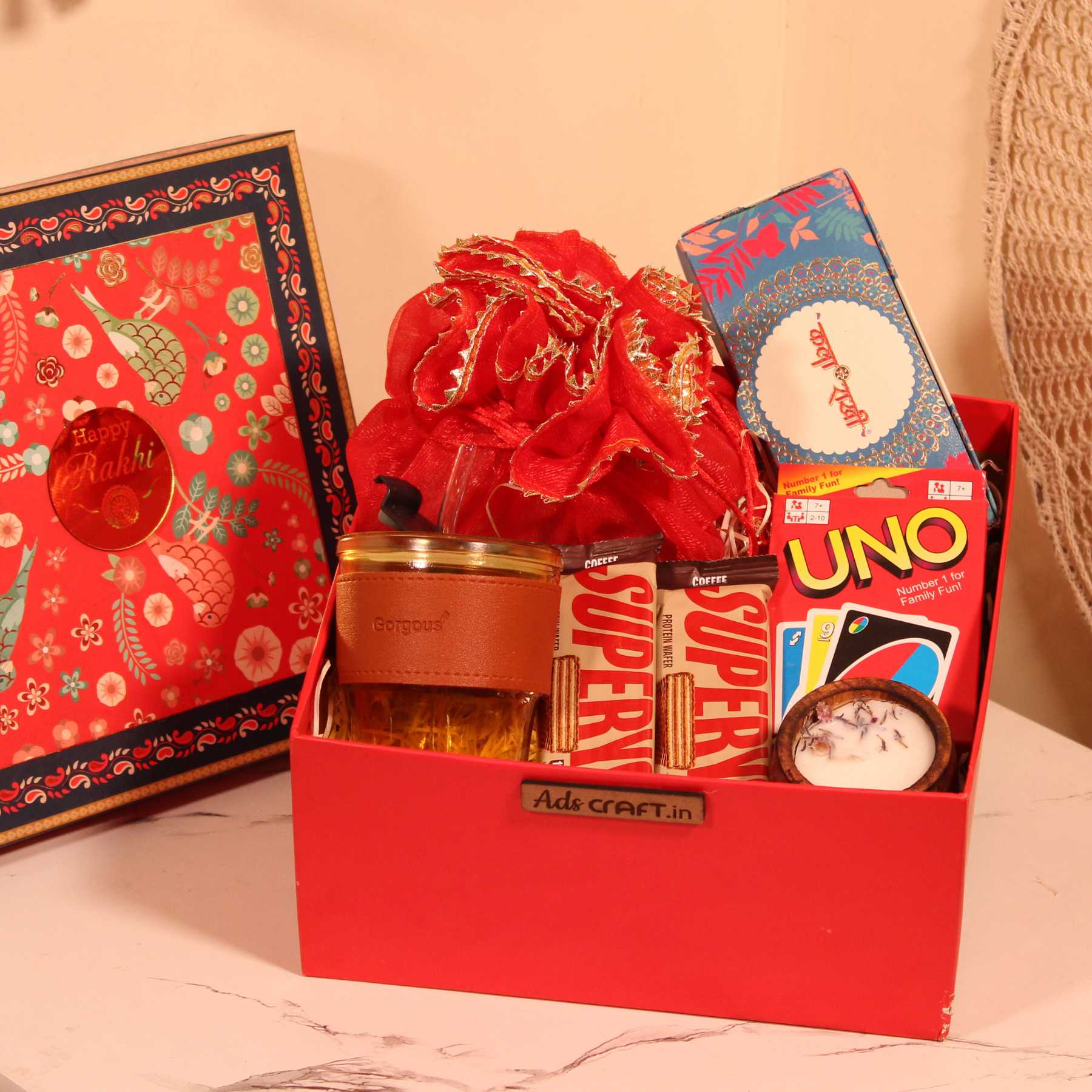 Rakhi gift hamper for brother in a red box with dry fruits, UNO cards, cookies, and glass sipper.